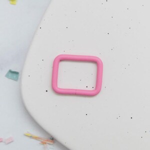 May include: A bright pink, rectangular metal buckle. The buckle is a solid, rounded rectangle shape with a small gap on one side. The buckle is set against a white background with small black specks.
