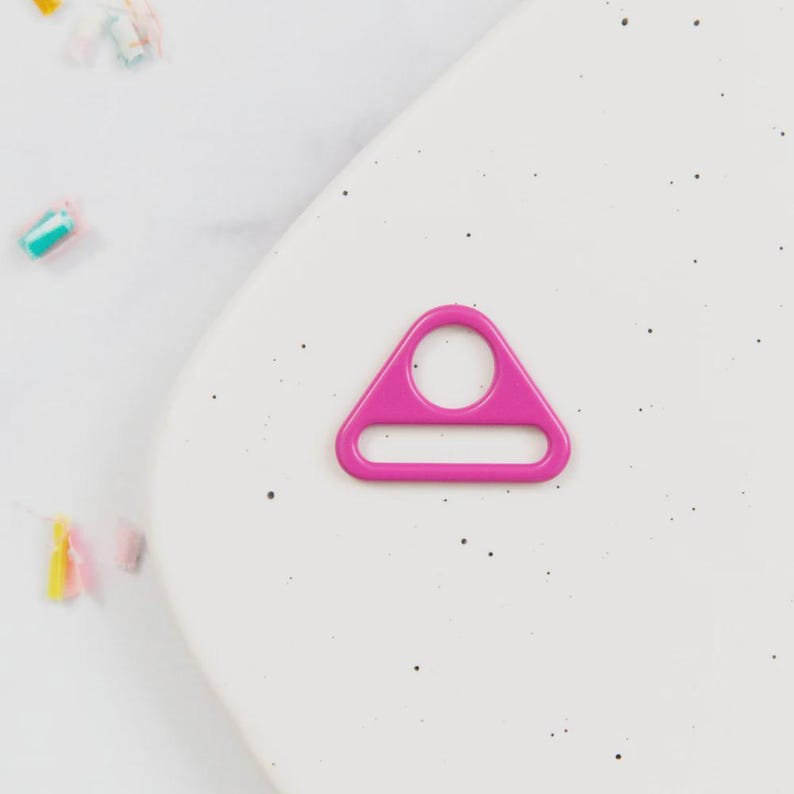 May include: A bright pink, triangular metal ring with a circular hole and a rectangular opening. The ring is set against a speckled white background, with colorful confetti scattered around the edges.