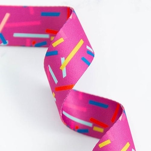 May include: A vibrant pink ribbon with a repeating pattern of colorful rectangular shapes in blue, yellow, and red. The ribbon is curled, showcasing its design and texture. The background is plain white.