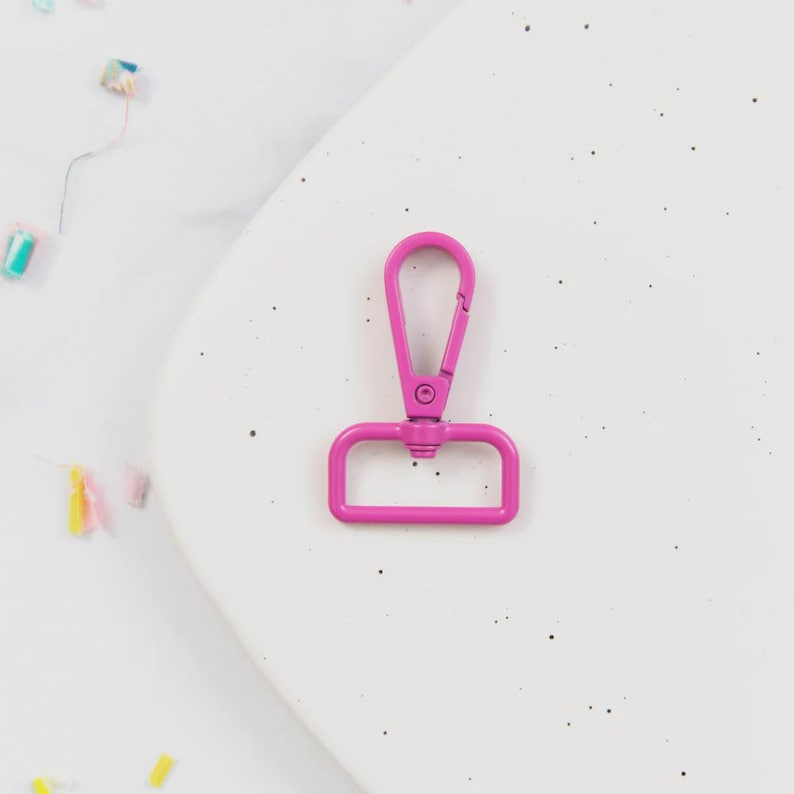 May include: A bright pink swivel clasp, a metal hardware component, is displayed against a white background. The clasp has a spring-loaded lever and a D-ring base, commonly used in crafting and bag making.