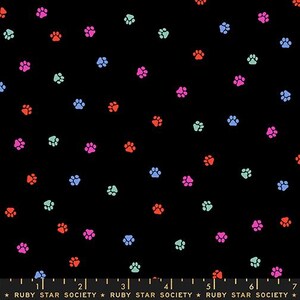 May include: A black fabric featuring a repeating pattern of small paw prints in vibrant shades of pink, orange, light blue, and mint green. A ruler at the bottom edge shows measurements in inches, with the text Ruby Star Society printed repeatedly.