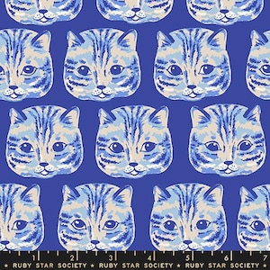 May include: A fabric pattern featuring repeating cat faces in shades of light blue, cream, and dark blue on a solid blue background. A black measuring tape with white markings and the text Ruby Star Society is visible along the bottom edge.