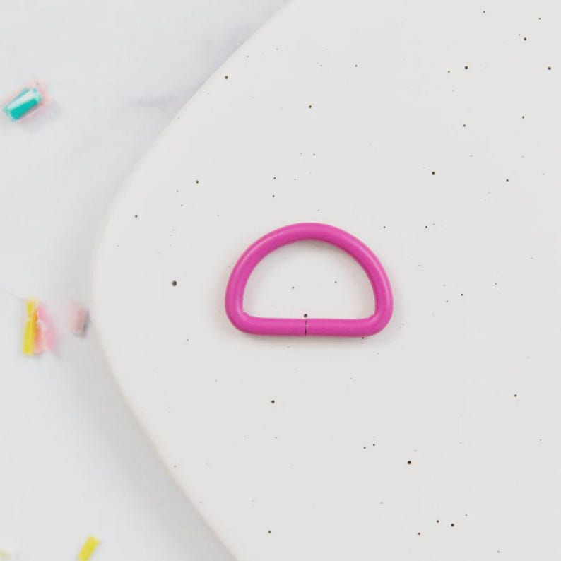 May include: A bright magenta-colored D-ring, a half-circle shape, lies on a speckled white surface. The D-ring is made of metal and has a smooth, even finish. This hardware component is used in various crafting and sewing projects.