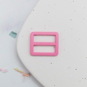 May include: A bright pink rectangular slider buckle with two horizontal bars. This plastic buckle is designed for use in various crafting and sewing projects, such as bag straps or clothing adjustments. The buckle is shown against a white background.