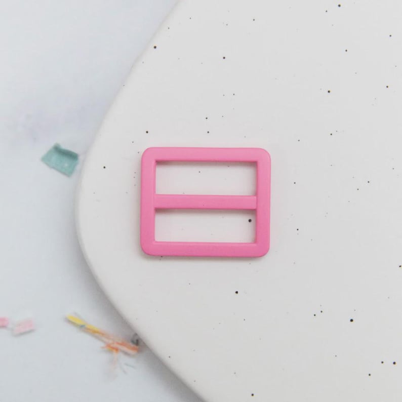 May include: A bright pink rectangular slider buckle with two horizontal bars. This plastic buckle is designed for use in various crafting and sewing projects, such as bag straps or clothing adjustments. The buckle is shown against a white background.