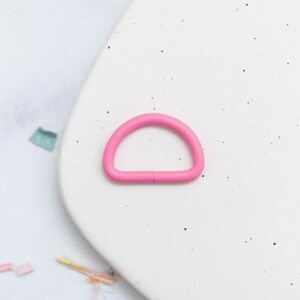 May include: A bright pink D-ring hardware component. The D-ring is made of a solid, smooth material and is set against a white background. The hardware is a half-circle shape with a small gap.
