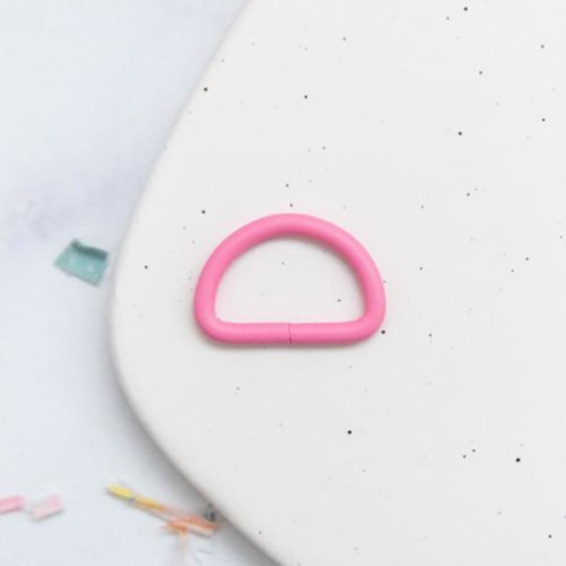 May include: A bright pink D-ring hardware component. The D-ring is made of a solid, smooth material and is set against a white background. The hardware is a half-circle shape with a small gap.