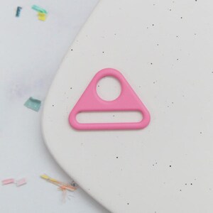 May include: A bright pink, triangular-shaped metal ring with a circular hole at the top and a rectangular opening at the bottom. The ring is set against a white background with small black speckles and colorful confetti.