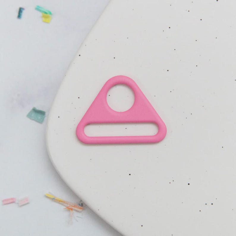 May include: A bright pink, triangular-shaped metal ring with a circular hole at the top and a rectangular opening at the bottom. The ring is set against a white background with small black speckles and colorful confetti.