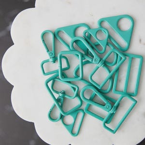 May include: A collection of teal-colored metal hardware pieces, including D-rings, rectangular rings, and swivel clasps. The hardware is arranged on a white, scalloped-edge surface. The pieces are new and unused.