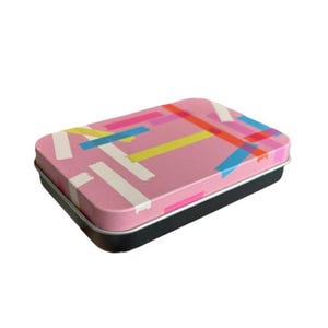 May include: A pink rectangular tin with a black lid. The tin has a colorful geometric pattern of overlapping stripes in pink, yellow, blue, and orange.