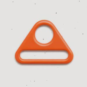 May include: A bright orange triangular strap adjuster buckle with a smooth, rounded finish. The hardware features a circular top opening and a wider, horizontal rectangular slot at the base, designed for threading webbing or fabric straps.