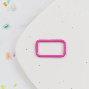 May include: A bright pink rectangular metal buckle. The buckle is a solid color and has rounded corners. The background is a speckled white surface with colorful confetti scattered around the buckle.