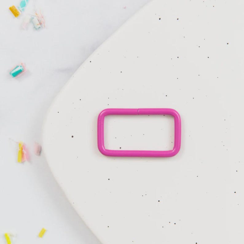 May include: A bright pink rectangular metal buckle. The buckle is a solid color and has rounded corners. The background is a speckled white surface with colorful confetti scattered around the buckle.