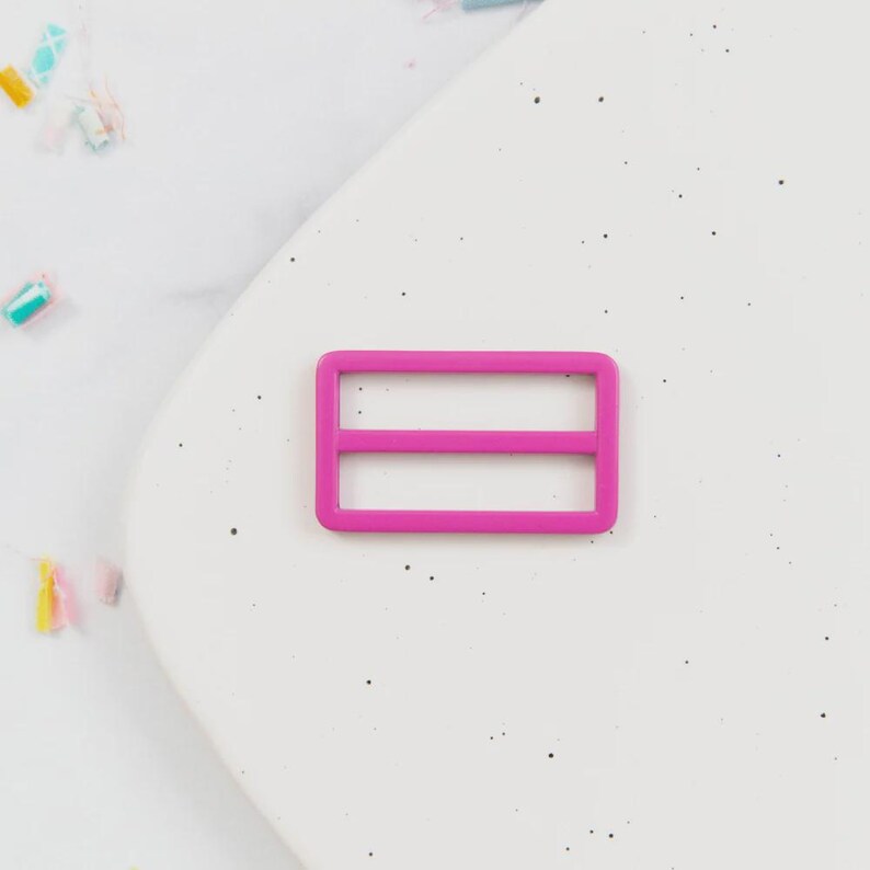 May include: A bright pink rectangular slider buckle with two horizontal bars. The buckle is made of plastic and is set against a white background. The slider buckle is a component used in various crafts and sewing projects.