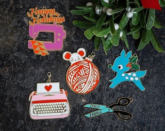 May include: Set of six gold-tone metal ornaments with a Christmas theme. The ornaments feature a sewing machine, a ball of yarn, a typewriter, a deer, a mouse, and a pair of scissors. The ornaments are decorated with red, white, blue, and green enamel paint. The ornaments have the text "Happy Holidays", "Only Fabric", and a red heart.
