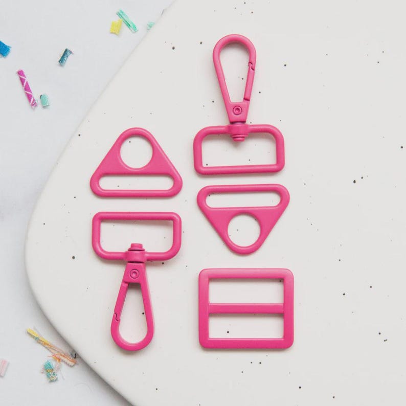 May include: A collection of bright pink metal hardware pieces, including a swivel clasp, a rectangular buckle, a triangle with a hole, a rectangle with a hole, and a D-ring. These components are likely for bag making or other craft projects.