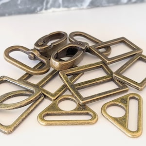 May include: An assortment of antique brass-colored metal hardware components, including D-rings, rectangular rings, triangular rings, and swivel clasps. The hardware pieces are scattered on a white surface, showcasing their various shapes and designs.