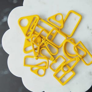 May include: A collection of bright yellow metal hardware pieces, including D-rings, rectangular rings, and swivel clasps. The hardware is scattered on a white, scalloped surface, creating a visually appealing arrangement. The pieces are likely for bag making or crafting.