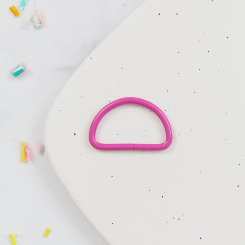 May include: A bright magenta D-ring, a half-circle shape, lies on a speckled white surface. The D-ring is made of metal and has a smooth finish. The background includes colorful confetti.