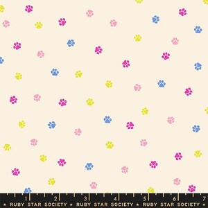May include: A cream-colored fabric featuring a repeating pattern of small paw prints in shades of bright pink, light pink, yellow, and blue. A black ruler with white markings and the text Ruby Star Society is visible at the bottom edge.