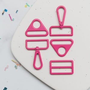 May include: A collection of bright pink hardware components, including a triangle, a rectangle, a square, and a clasp. The pieces are arranged on a white speckled surface, suggesting a crafting or DIY theme.