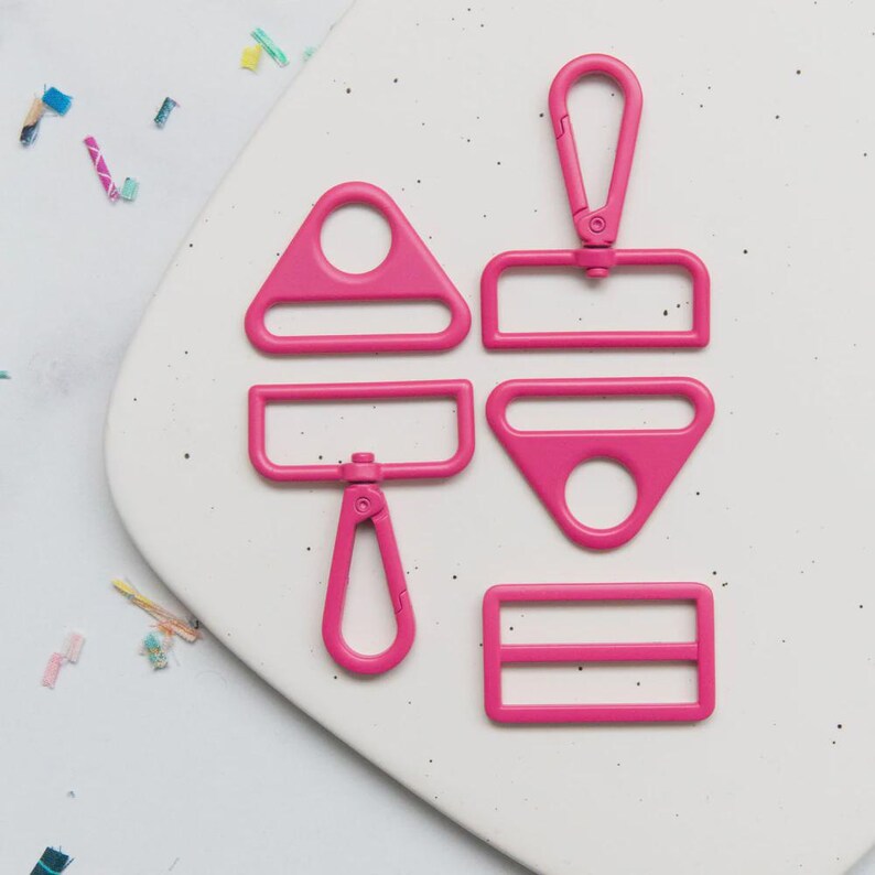 May include: A collection of bright pink hardware components, including a triangle, a rectangle, a square, and a clasp. The pieces are arranged on a white speckled surface, suggesting a crafting or DIY theme.