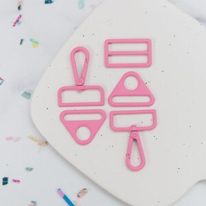 May include: A collection of pink metal hardware pieces, including a rectangular buckle, a D-ring, a triangle-shaped piece, and two swivel clasps. The hardware is arranged on a white, speckled surface.