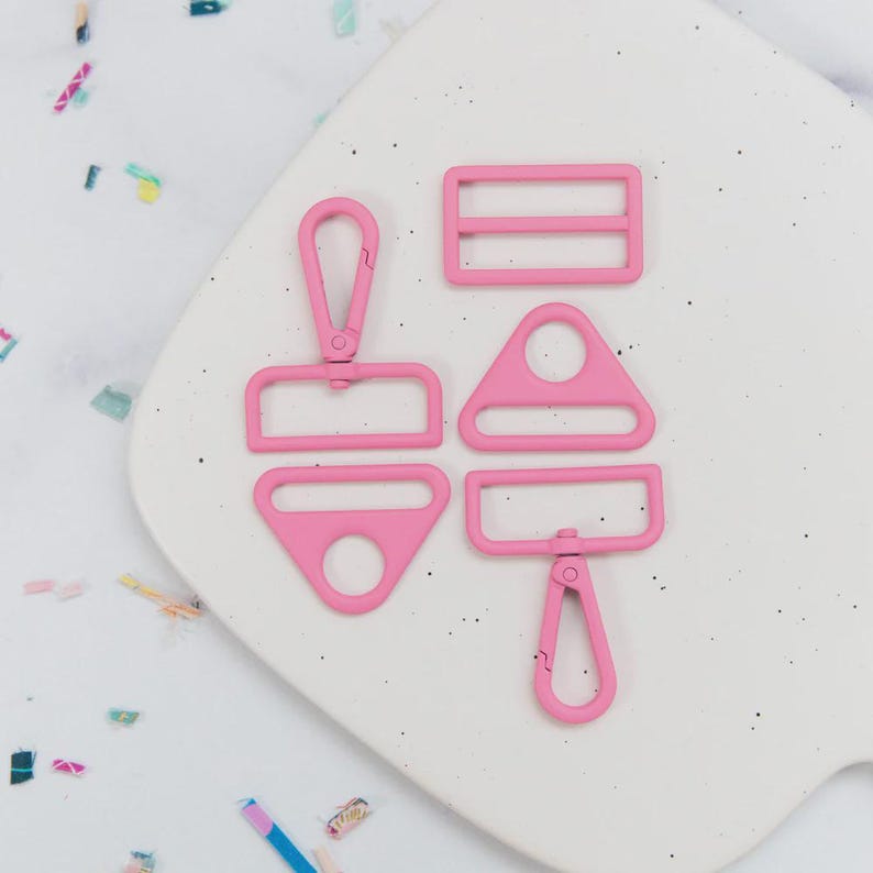 May include: A collection of pink metal hardware pieces, including a rectangular buckle, a D-ring, a triangle-shaped piece, and two swivel clasps. The hardware is arranged on a white, speckled surface.