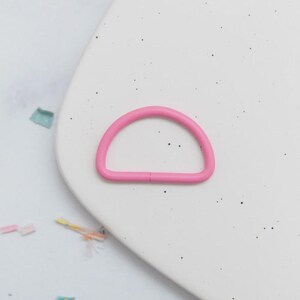 May include: A bright pink D-ring, a half-circle shape, sits on a white speckled surface. The D-ring is made of a solid material and has a small gap where the ends meet. Small colorful confetti pieces are scattered around the ring.