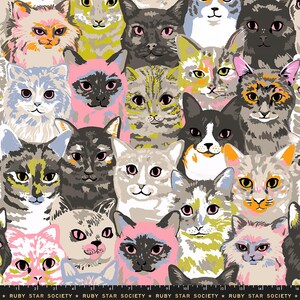 May include: A fabric print featuring a repeating pattern of various cat faces in diverse colors including pink, gray, yellow, and black. The cats have distinct facial markings and expressions. The bottom edge displays the text RUBY STAR SOCIETY.