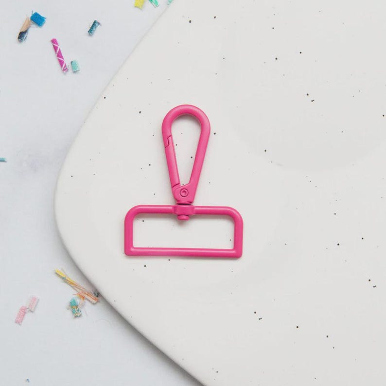 May include: A bright pink metal swivel clasp, designed for use with bags or straps. The clasp features a rectangular base and a curved top with a spring-loaded opening. The hardware is set against a white background with confetti.