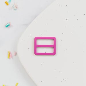 May include: A bright pink rectangular slider buckle with a horizontal bar in the middle. The buckle is set against a white background with small black specks and colorful confetti.
