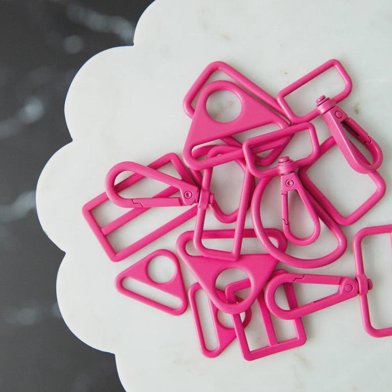 May include: A collection of bright pink metal swivel clasps and D-rings in various shapes. The clasps have a spring-loaded mechanism for easy attachment. The items are scattered on a white, scalloped-edge surface.