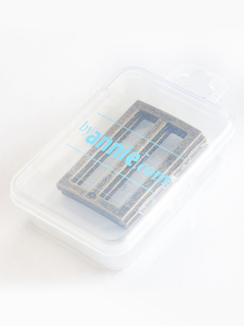 Puede incluir: Una hebilla de metal de color dorado con forma rectangular y tres barras horizontales. La hebilla est&aacute; en un paquete de pl&aacute;stico transparente con el texto "by annie.com" impreso en el lateral.