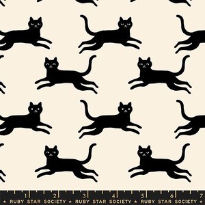 May include: A cream-colored fabric featuring a repeating pattern of black cats in a running pose. A black ruler at the bottom edge displays measurements in inches, with the text Ruby Star Society printed repeatedly along the length of the ruler.