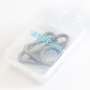 Puede incluir: Dos llaveros de metal de color dorado antiguo en una caja de almacenamiento de pl&aacute;stico transparente. La caja tiene una etiqueta que dice "by annie.com".
