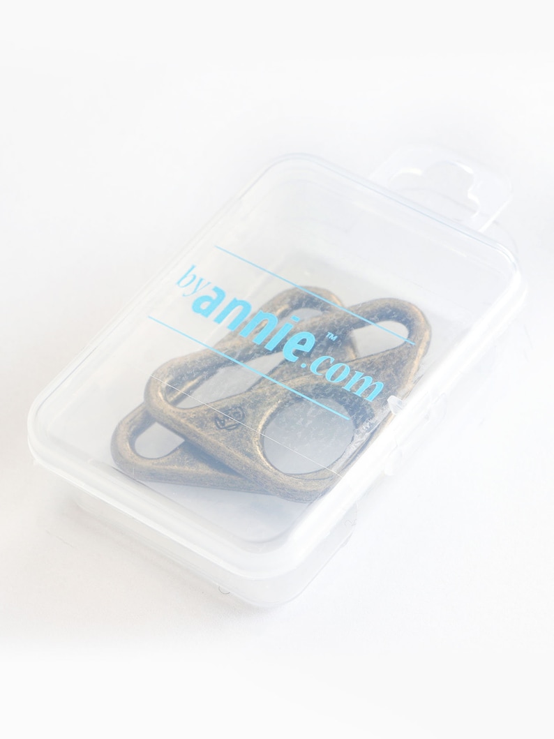 Puede incluir: Dos llaveros de metal de color dorado antiguo en una caja de almacenamiento de pl&aacute;stico transparente. La caja tiene una etiqueta que dice "by annie.com".