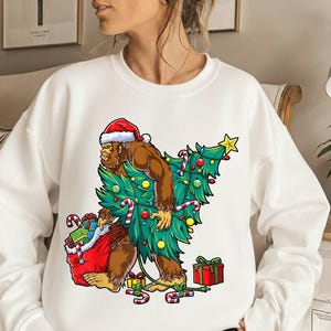 Könnte beinhalten: Weißes Sweatshirt mit einem Bigfoot-Charakter, der einen Weihnachtsmannhut trägt und einen Weihnachtsbaum trägt. Der Bigfoot trägt auch einen roten Geschenkesack und ist von Zuckerstangen und Geschenken umgeben. Ein festliches Weihnachtsdesign.