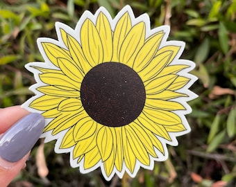 May include: A die-cut sticker featuring a bright yellow sunflower with a dark brown center. The sunflower has detailed petals and a white border. The sticker is held against a background of green foliage.