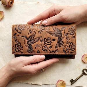 May include: A rectangular brown leather wallet featuring an embossed design of two hummingbirds in flight surrounded by detailed rose blossoms and swirling vine patterns. The leather has a matte finish with dark brown accents in the engravings.