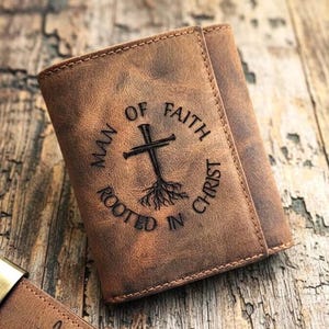 May include: A brown leather tri-fold wallet featuring an embossed design of a cross with roots at the base. The text MAN OF FAITH ROOTED IN CHRIST is engraved in a circular pattern around the cross. A matching brown leather keychain is visible.