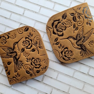 Puede incluir: Dos carteras de cuero color canela con un dise&ntilde;o de colibr&iacute; y rosas. Las carteras tienen una parte superior curvada y est&aacute;n decoradas con un colibr&iacute;, rosas y hojas en relieve. El fondo es una pared de ladrillos blancos.