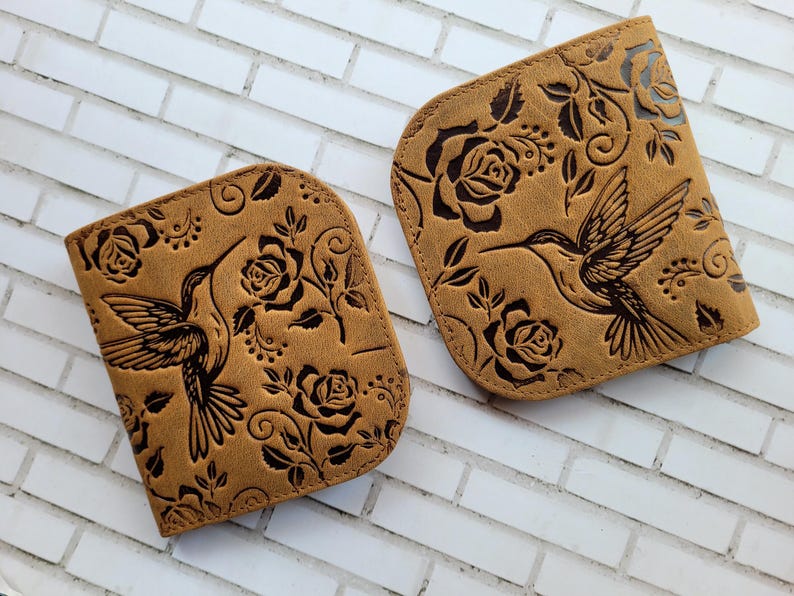Puede incluir: Dos carteras de cuero color canela con un dise&ntilde;o de colibr&iacute; y rosas. Las carteras tienen una parte superior curvada y est&aacute;n decoradas con un colibr&iacute;, rosas y hojas en relieve. El fondo es una pared de ladrillos blancos.