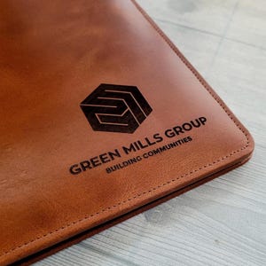 May include: A brown leather folder with a logo embossed on the front. The logo is a geometric shape with the text "GREEN MILLS GROUP BUILDING COMMUNITIES" below it.