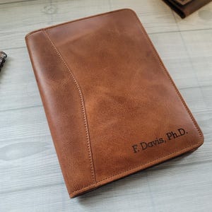 Könnte beinhalten: Ein braunes Leder-Portfolio mit dem eingravierten Text "F. Davis, Ph.D." in der unteren rechten Ecke. Das Portfolio ist geschlossen und hat ein genähtes Design entlang der linken Seite. Ein Spiralnotizblock und ein Stift befinden sich im Hintergrund.