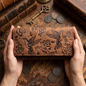 May include: A rectangular brown leather wallet featuring an embossed design of two hummingbirds in flight surrounded by detailed rose blossoms and swirling vine patterns across the front surface of the accessory.