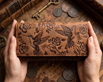 Puede incluir: Una cartera rectangular de cuero marrón con un diseño en relieve de dos colibríes en vuelo rodeados de detalladas flores de rosa y patrones de enredaderas en espiral en la superficie frontal del accesorio.