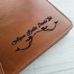 May include: A brown leather folder with the text "Aqua Bella Pools Inc." and a mermaid design embossed on the front.