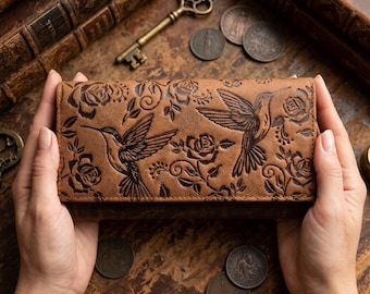 Puede incluir: Una cartera rectangular de cuero marrón con un diseño en relieve de dos colibríes en vuelo rodeados de detalladas flores de rosa y patrones de enredaderas en espiral en la superficie frontal del accesorio.